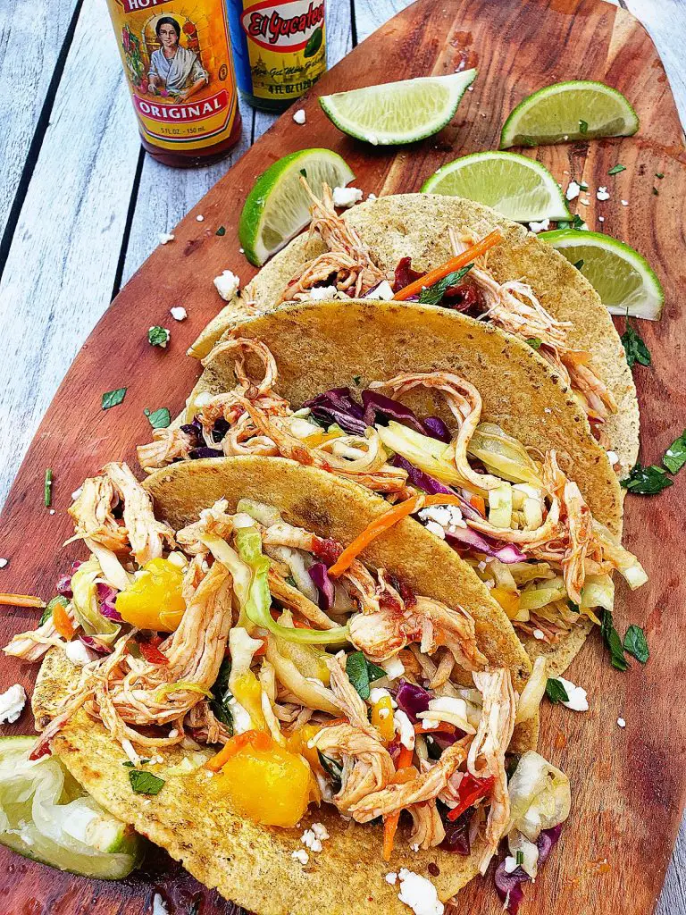 overhead side view of three chipotle chicken tacos with mango lime slaw on a wooden serving board served with lime wedges and hot sauce bottles