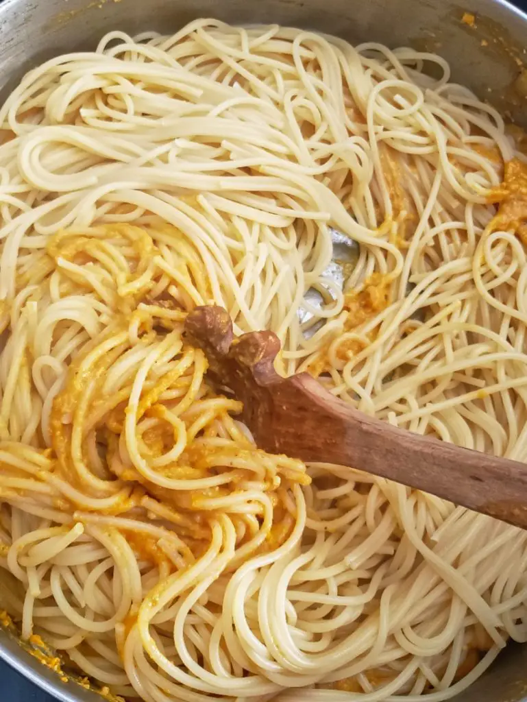 overhead image of spaghetti being combined with butternut squash sauce in a stainless steel skillet by a wooden pasta spoon
