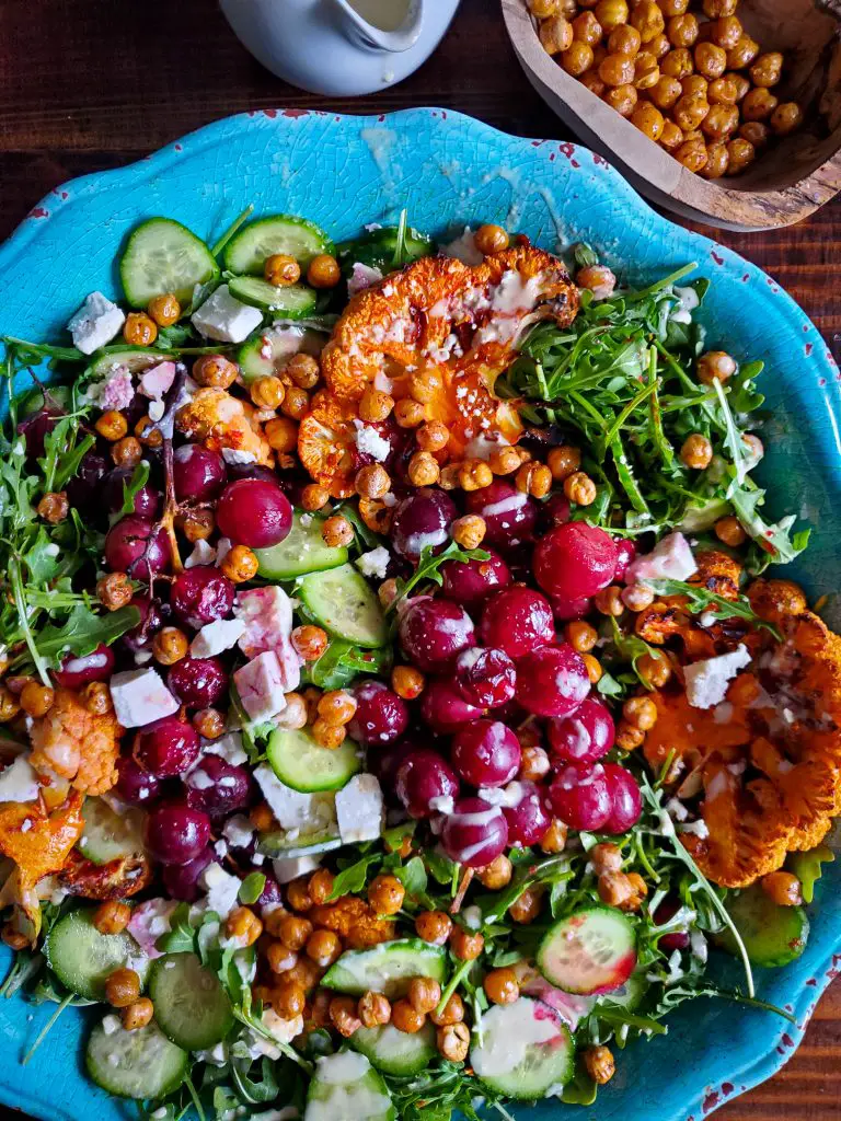 overhead image of middle eastern salad with harissa cauliflower roasted grapes and lemon tahini dressing next to wooden bowl of crispy chickpeas