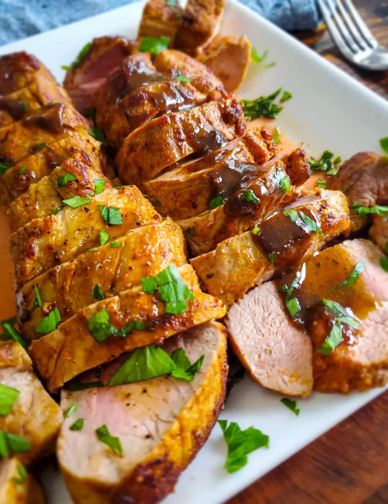 Side angel view of sliced pork tenderloin on white plate with sauce and chopped parsley on top