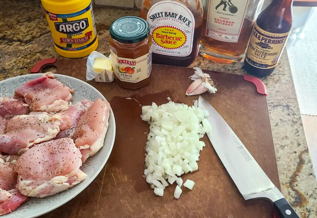 image of ingredients for peach and bourbon bbq chicken; chopped onion, garlic, chicken thighs, butter, peach preserves, bottle of bbq sauce, bottle of bourbon, and bottle of Worcestershire sauce