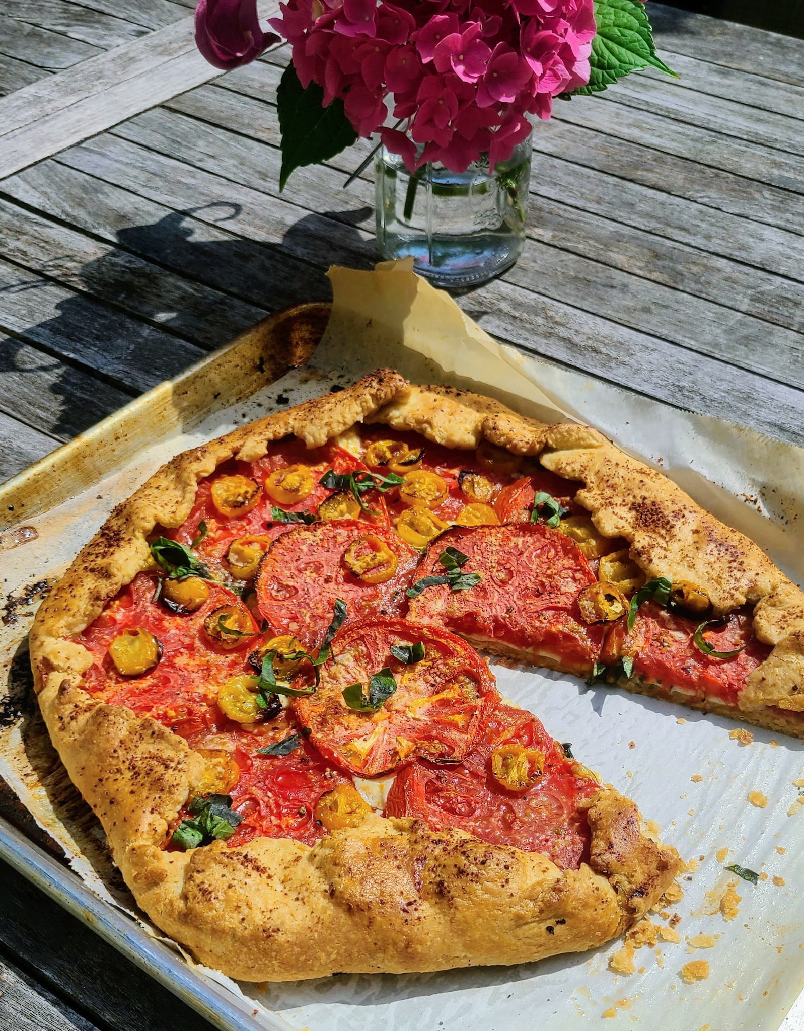 summer heirloom tomato galette sitting on sheet pan on an outdoor wooden table next to a vase of pink hydrangeas
