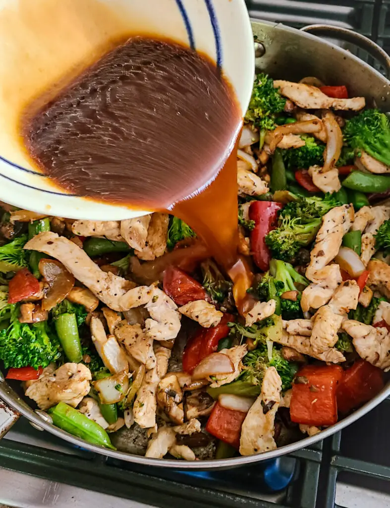 image of chicken and vegetable stir-fry in skillet with an Asian flavored brown sauce being poured over the top from a mixing bowl