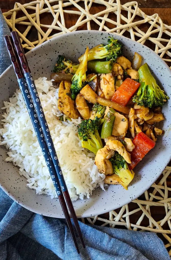overhead image displaying a bowl of white rice and chicken and vegetable stir fry. chopsticks are place along the bowl as well as a blue napkin and decorative placemat for aesteic effect