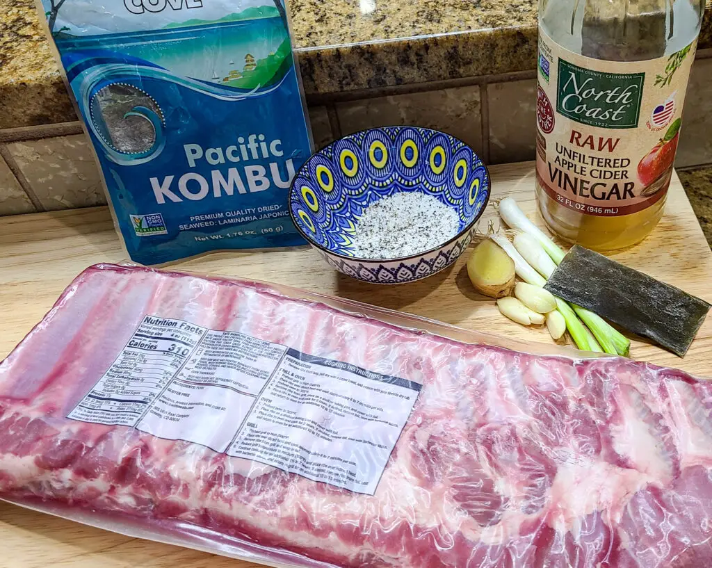 image of bag of kombu, bowl of salt and pepper, bottle of apple cider vinegar, scallions, knob of ginger, cloves of garlic with are ingredients added to instant pot to pressure cook spare ribs