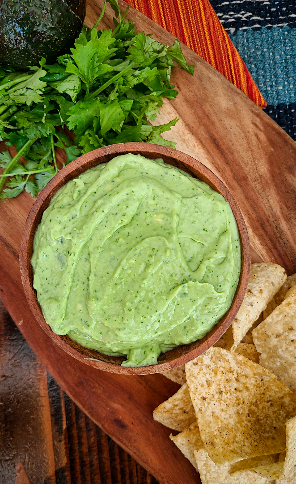 overhead image of whipped guacamole dip in a wooden bowl on a wooden tray with a handful of cilantro to the left and a handful of chips to the right of the bowl