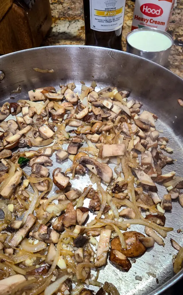 Image of sautéed baby Bella mushrooms and onions in skillet next to bottle of sherry wine and cup of cream