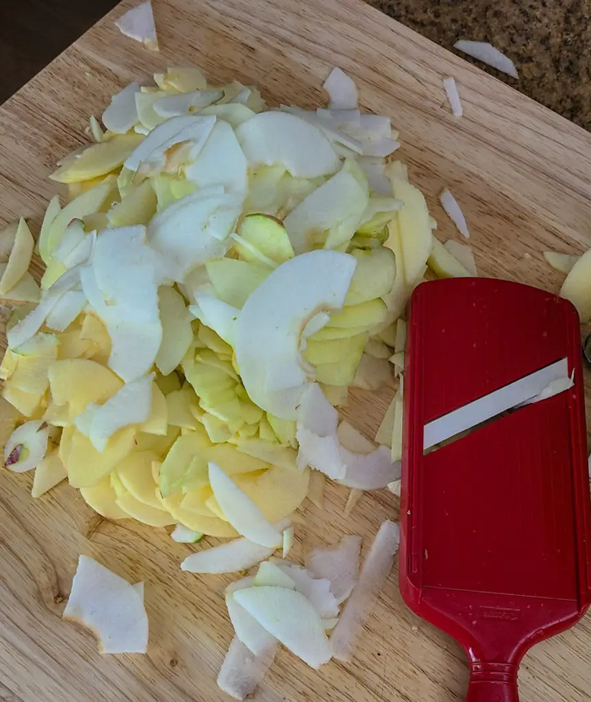 image of pile of thinly sliced apples next to mandoline on wooden cutting board