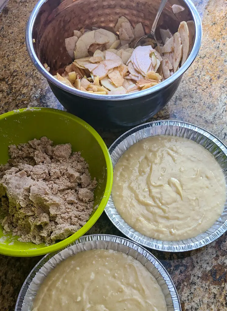 image of assembling crumb cake with bowl of apples, bowl of cinnamon crumbs, and two cake pans with pour batter in them