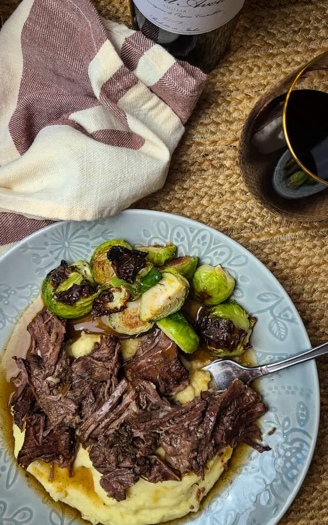 plate of shredded instant pot chuck roast with gravy over mashed potatoes with side of roasted brussels sprouts on dining table with red wine.