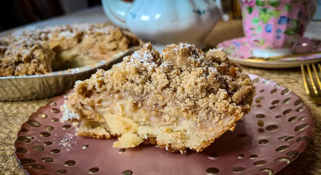 image of wedge of apple cinnamon crumb cake on pink plate with tea cup and tea pot in background