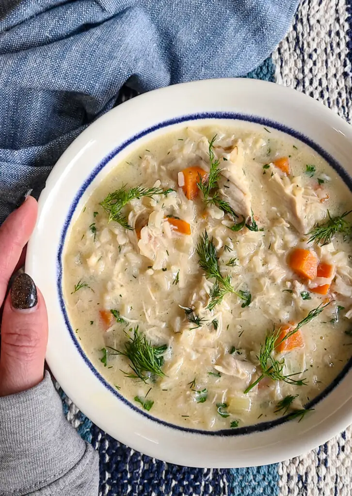 bowl of avgolemono soup with hand holding the side of bowl next to blue napkin