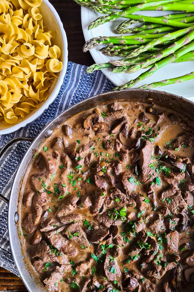beef stroganoff next to bowl of egg noodlles and tray of asparagus
