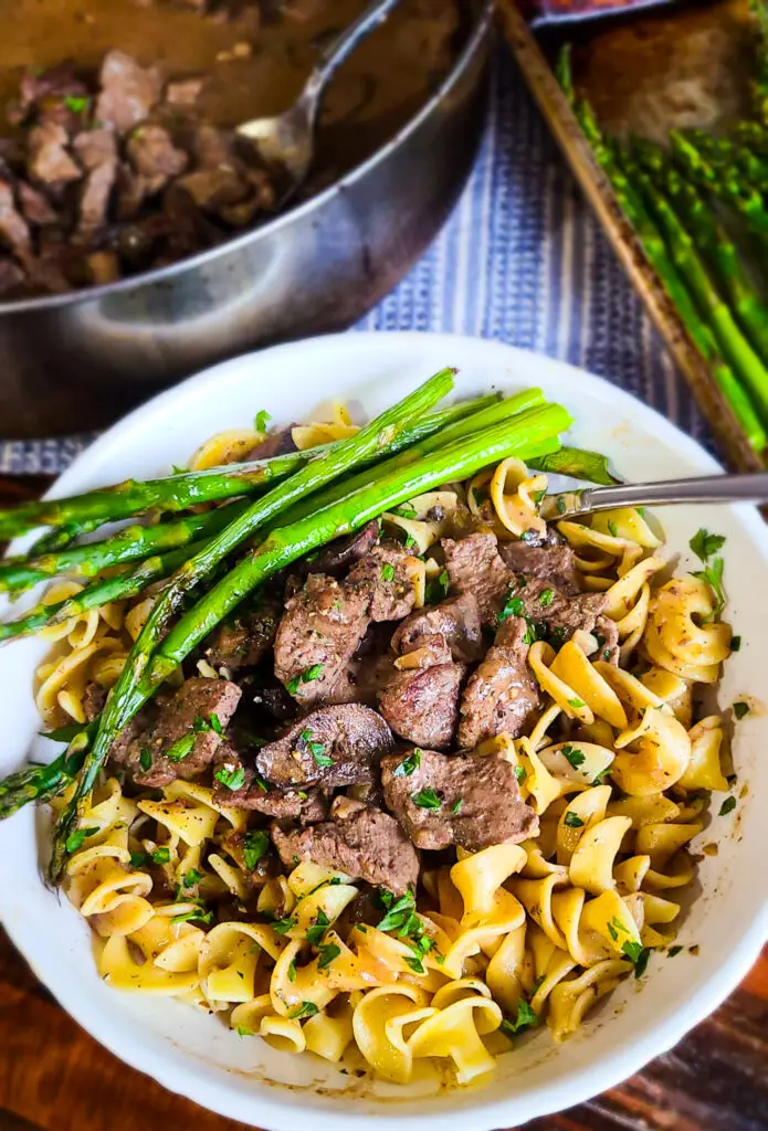 overhead image of beef stroganoff over egg noodles in white shallow bowl with serving of asparagus spears on the side