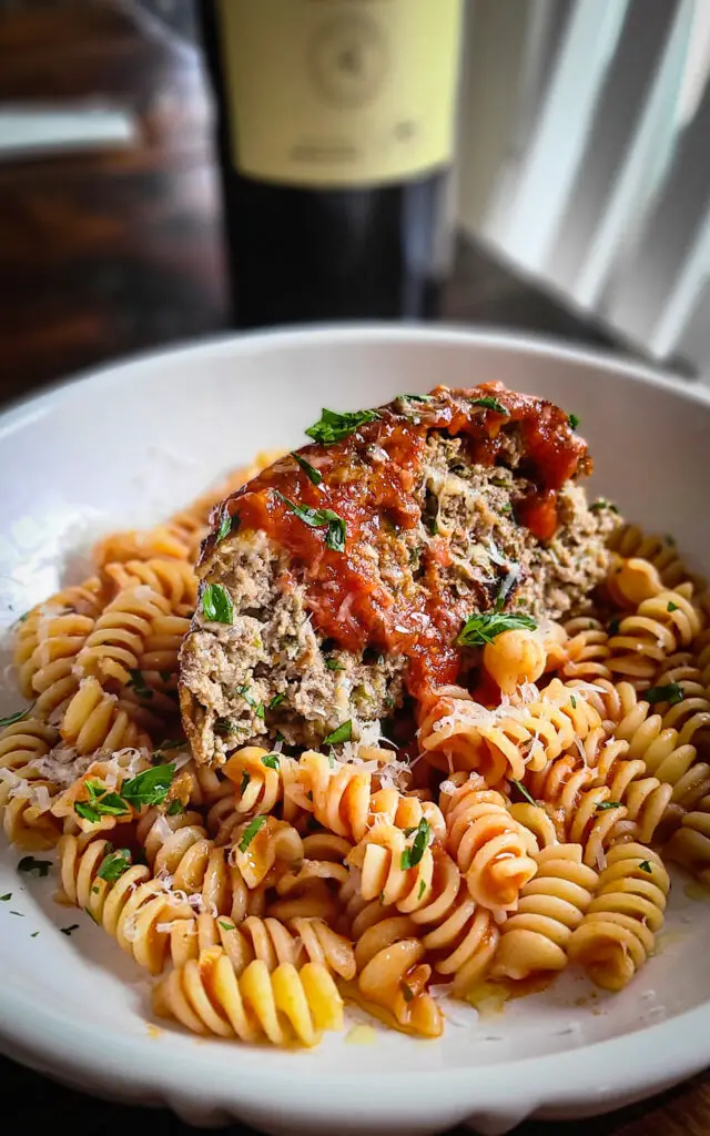 image of plate of Italian style meatloaf served over pasta with marinara sauce next to a bottle of red wine 