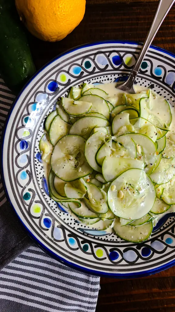 vertical image of cucumber salad with creamy italian dressing next to striped napkin and whole lemon and cumcumber