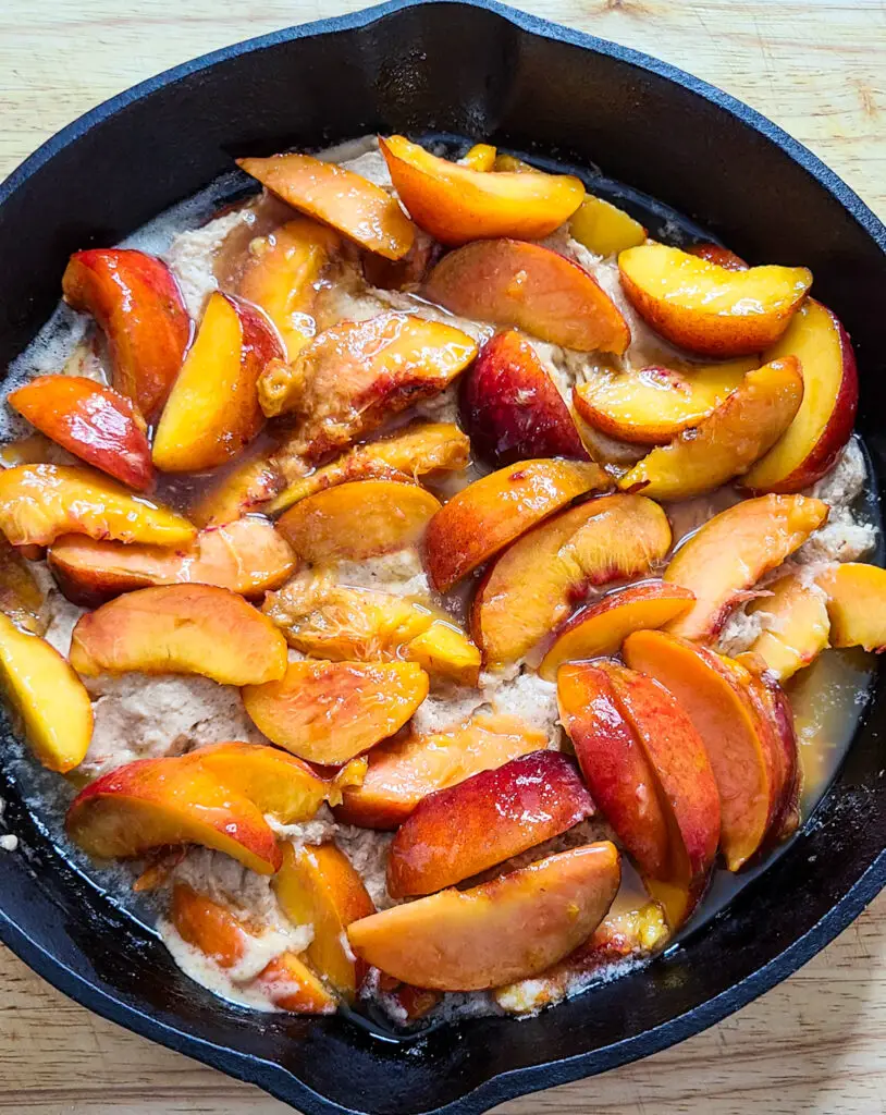 sliced peaches with cobbler batter and melted butter ready to bake in cast iron skillet