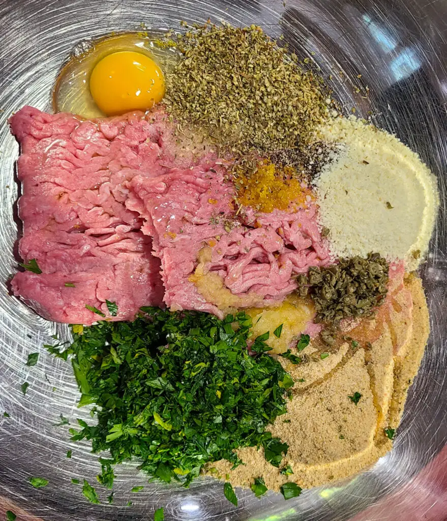 stainless steel mixing bowl with unmixed ingredients to make chicken piccata meatballs: ground chicken, egg, parsley, breadcrumbs, garlic, lemon capers, grated cheese