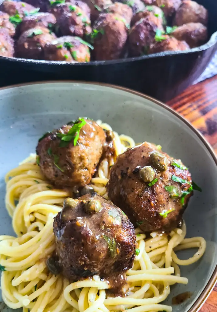 chicken piccata meatballs over a bed of spaghetti on dinner plate set next to cast iron pan containing more meatballs