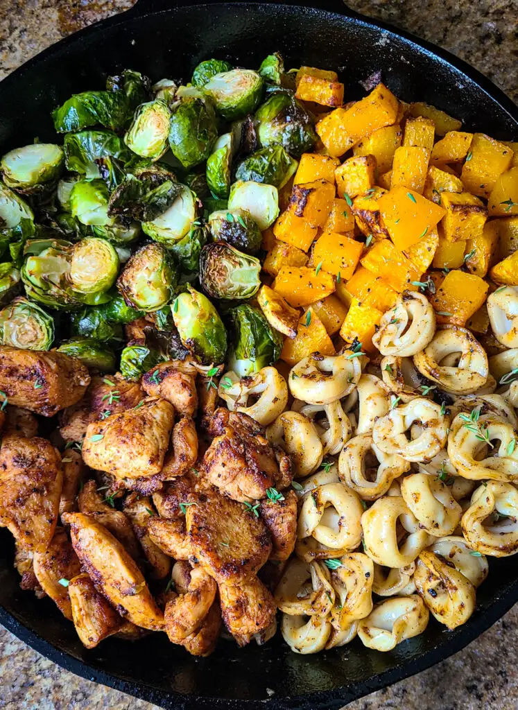 large cast iron pan with seared chicken slices, tortellini, roasted brussels sprouts and butternut squash separated in four quadrants in the pan