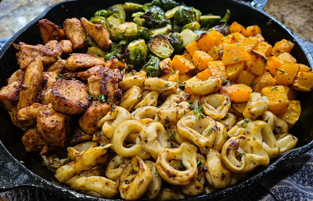 large cast iron pan with seared chicken slices, tortellini, roasted brussels sprouts and butternut squash separated in four quadrants in the pan