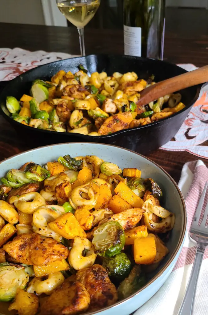 tablescape of bowl of Chicken and Tortellini with Autumn Roasted Vegetables in a serving bowl next to fork. a large cast iron pan containing more of the dinner next to a poured glass of white wine in in the background of photo.