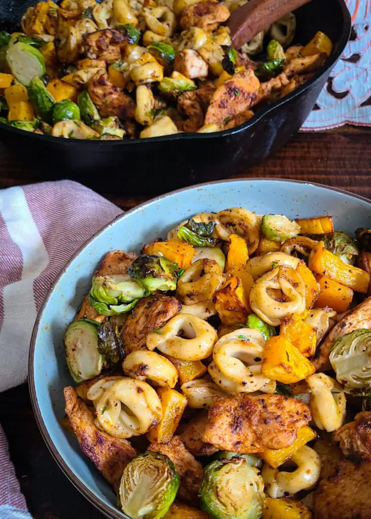 tablescape of bowl of Chicken and Tortellini with Autumn Roasted Vegetables in a serving bowl. a large cast iron pan containing more of the dinner in the background of photo.