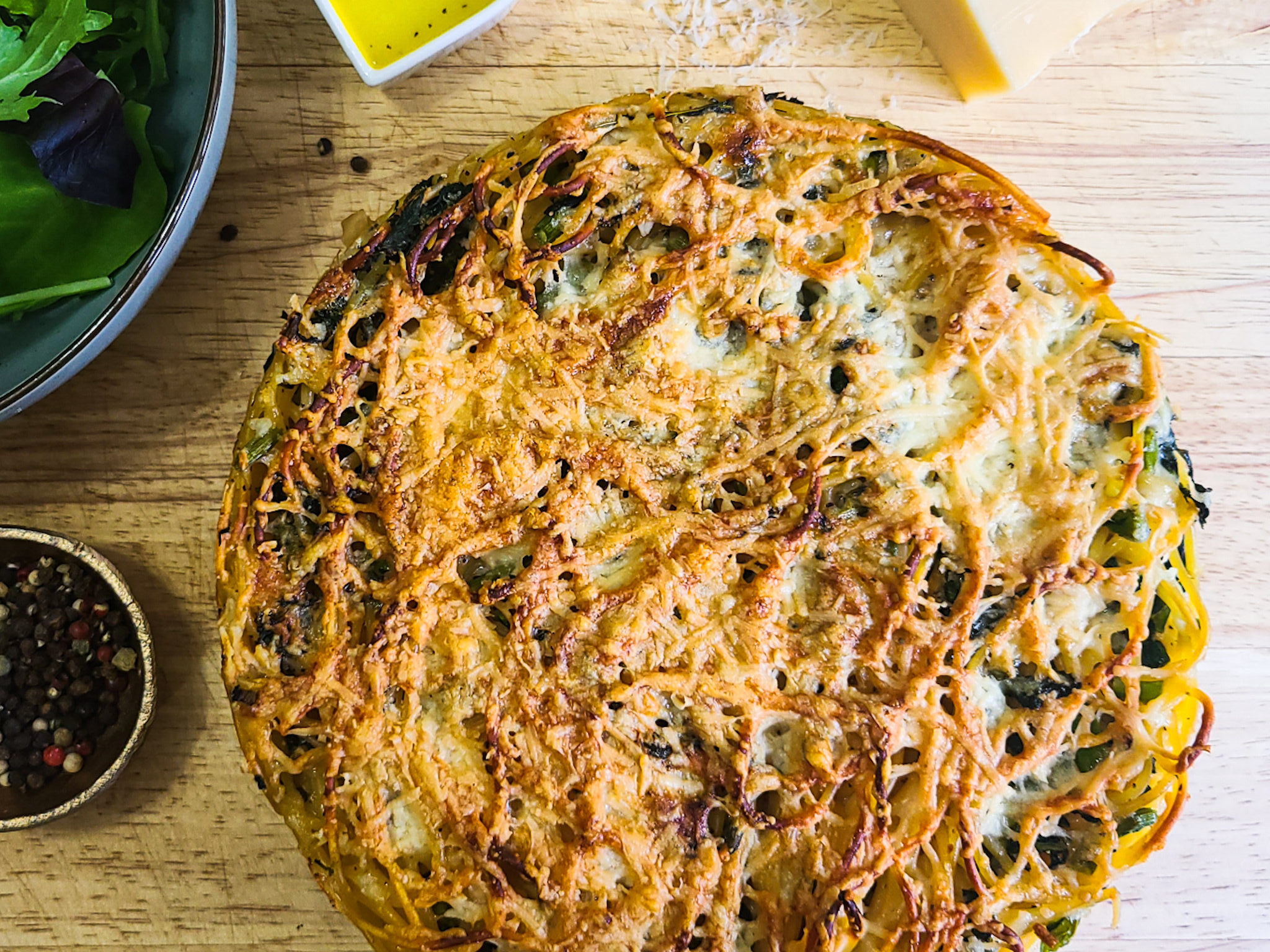 overhead image of cooked spaghetti frittata pie on a wooden board with bowl of peppercorns and salad used as visual props. 