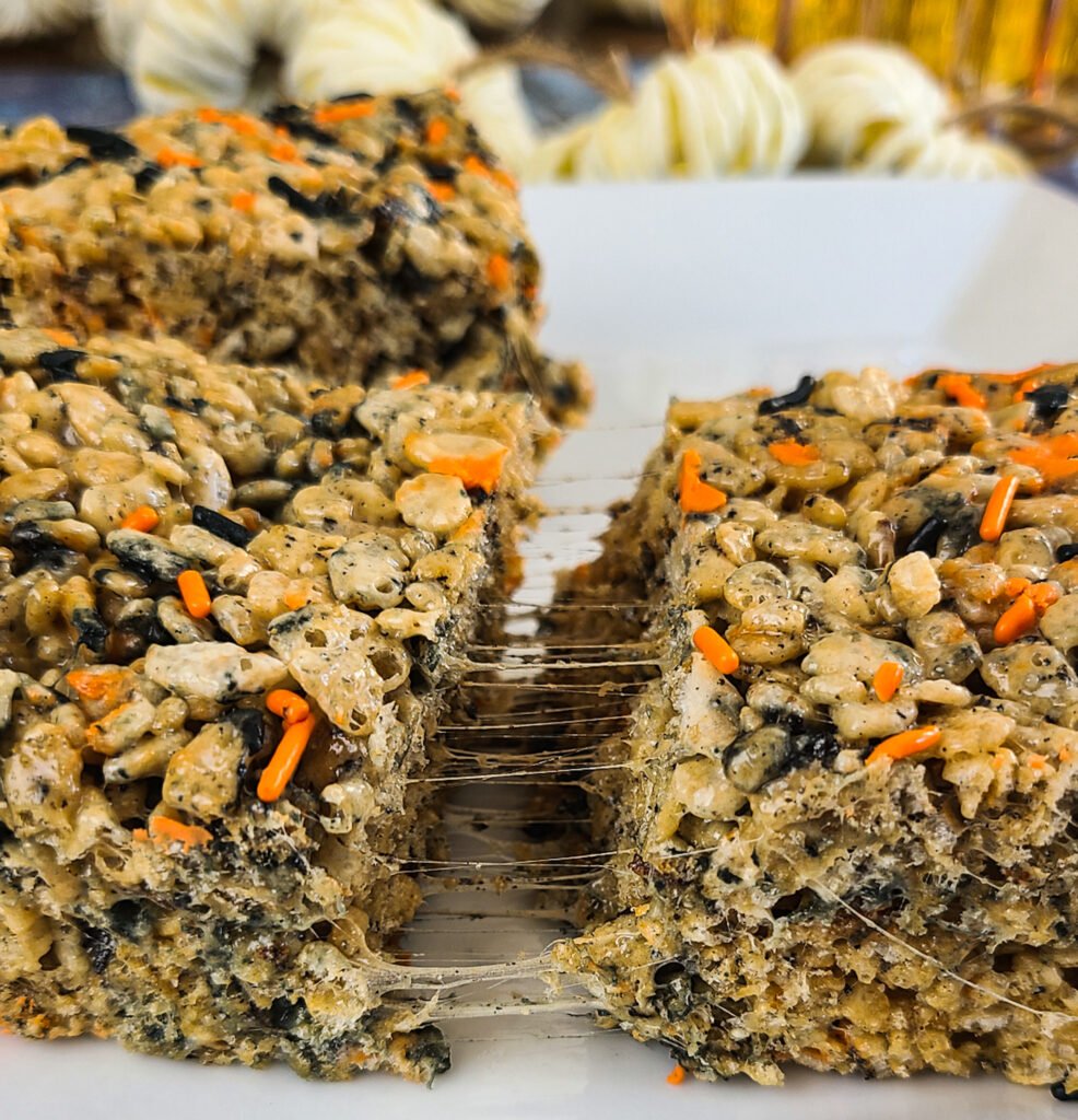 upclose image of gooey rice krispie treats with halloween colored sprinkles torn in half to show the melted marshmallows