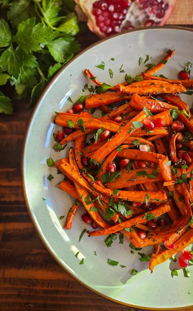 portrait overhead image of platter with oven roasted carrots cut into quarters. Carrots are drizzled with pomegranate molasses and garnished with chopped parsley and pomegranate seeds. Platter sit next to bunch of fresh parsley and halved pomegranate